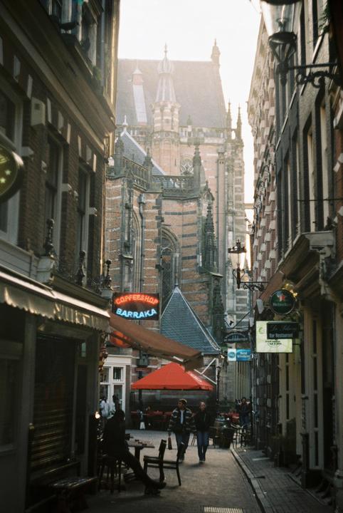 A narrow street in Amsterdam is lined with shops and restaurants, with a large historic building visible in the background.