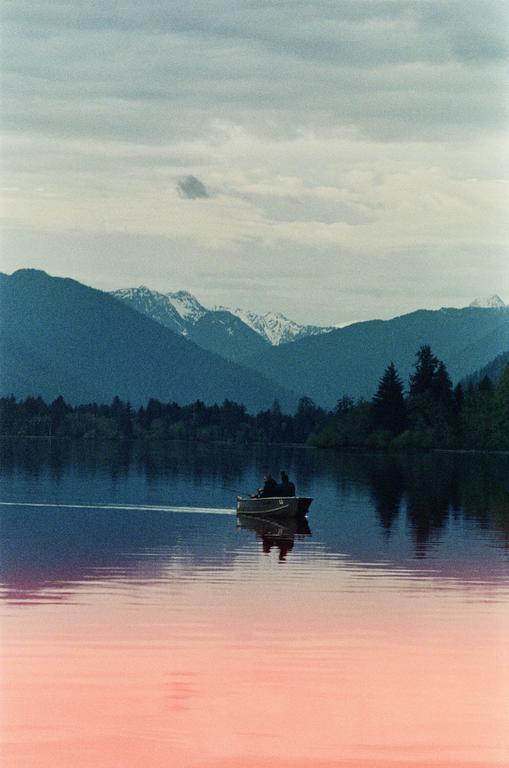 Two people in a small boat move across a lake with mountains and trees in the background.