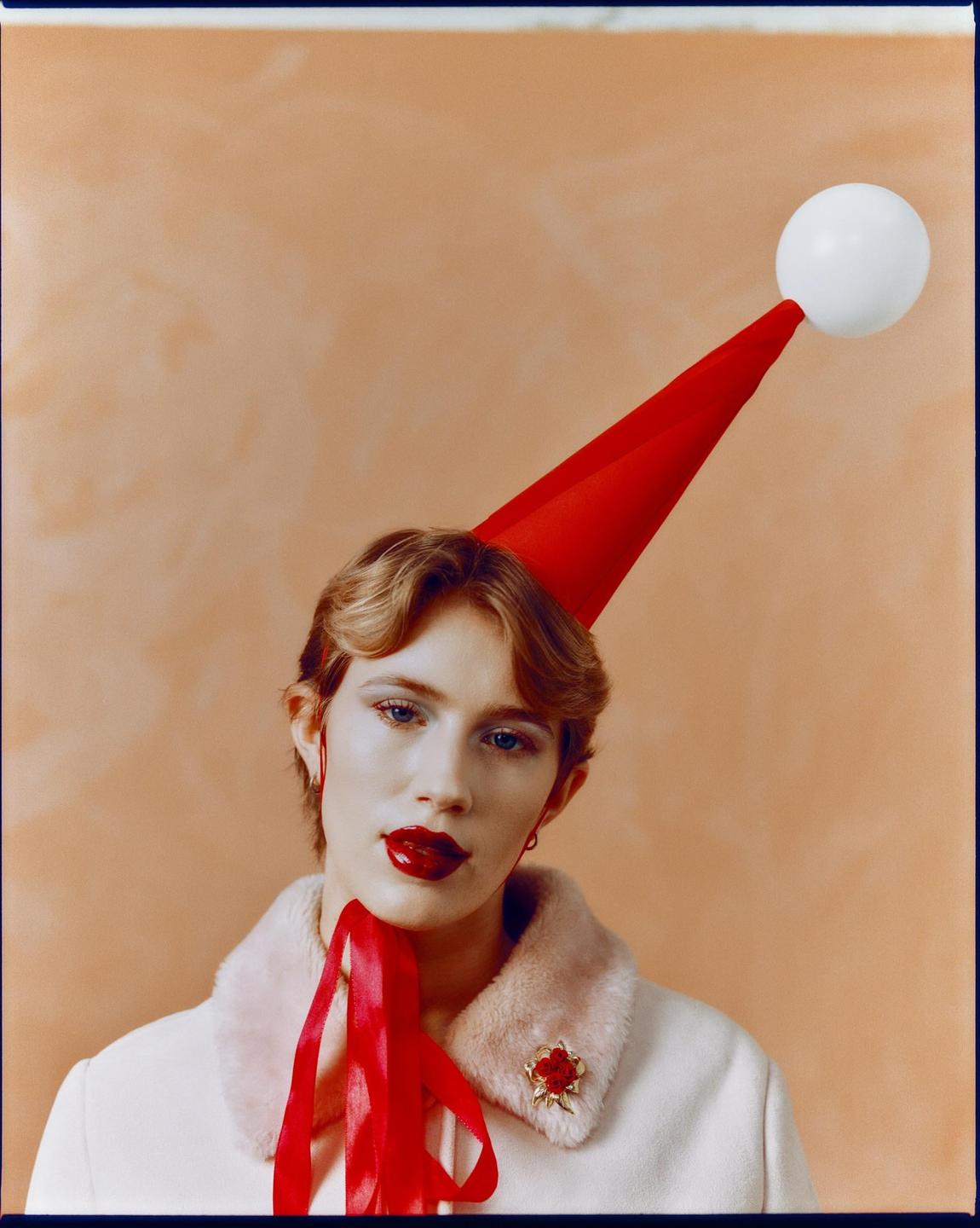 A woman with red lipstick and a red party hat with a white ball on top looks directly at the viewer.