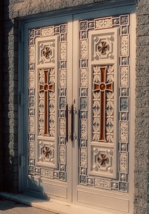 A white ornate door with crosses and floral patterns is set into a brick wall.