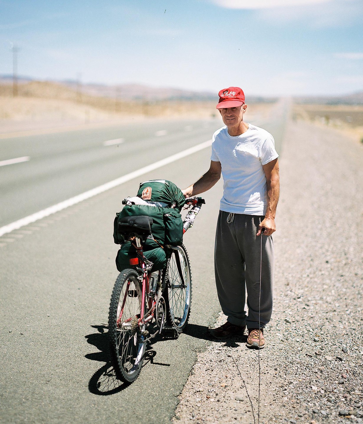 A man in a red cap stands next to a bicycle loaded with gear on the side of a long, empty road.