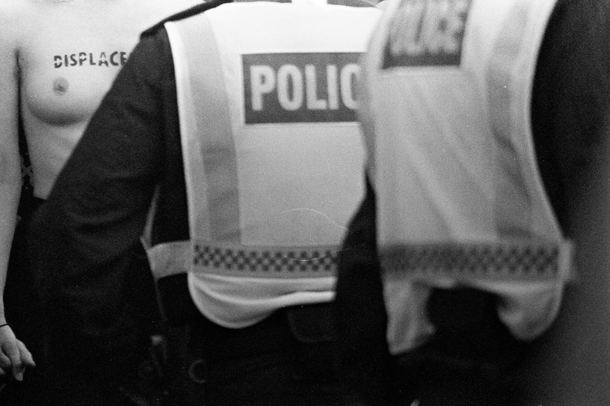 A woman with "DISPLACE" written on her chest stands next to two police officers.