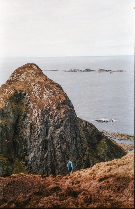 A person in a blue jacket stands on a grassy hill overlooking a rocky mountain and the ocean with a lighthouse in the distance.