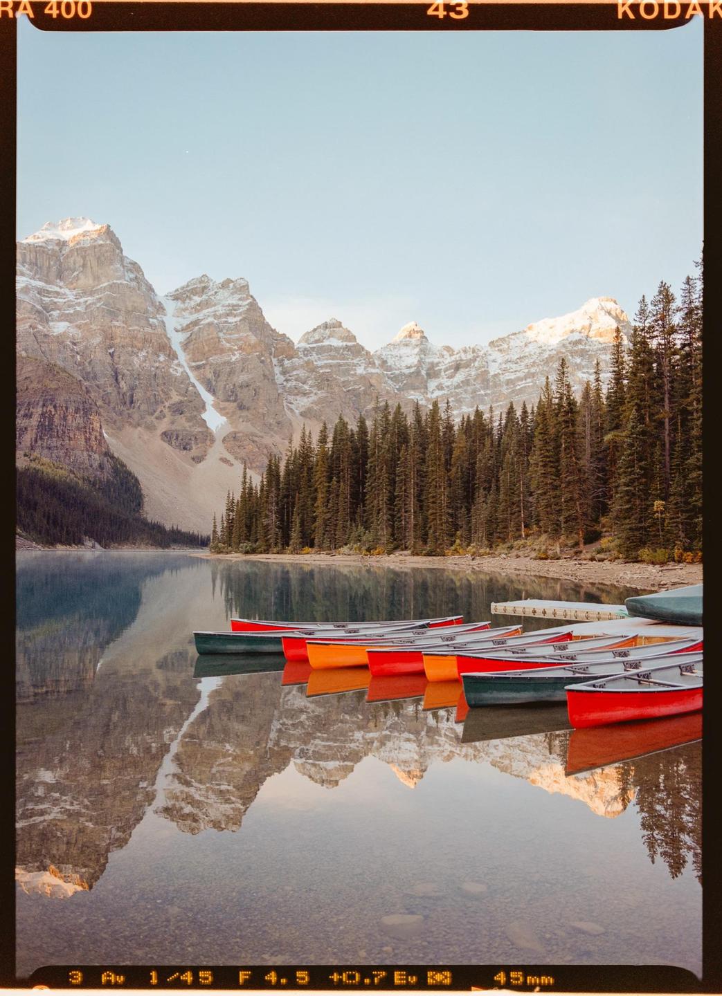 Colorful canoes are lined up on the shore of a lake with mountains and a forest in the background.