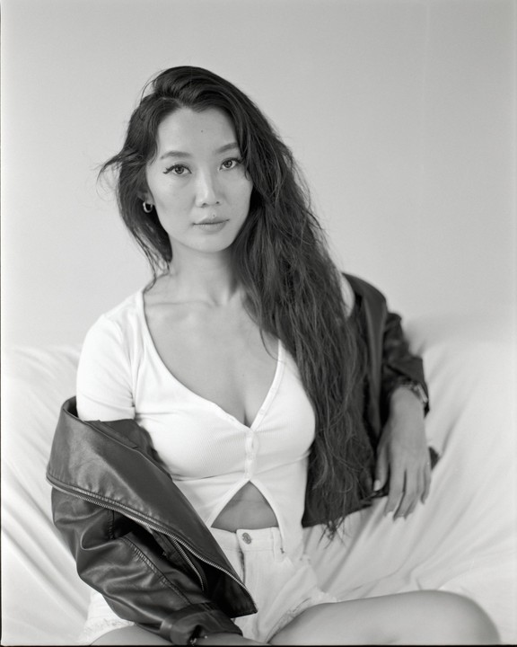 A woman with long hair and a leather jacket sits on a bed.