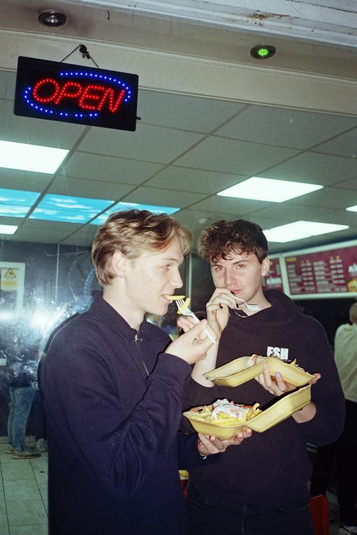 Two young men eat food from takeaway containers in a kebab shop under an illuminated "OPEN" sign.