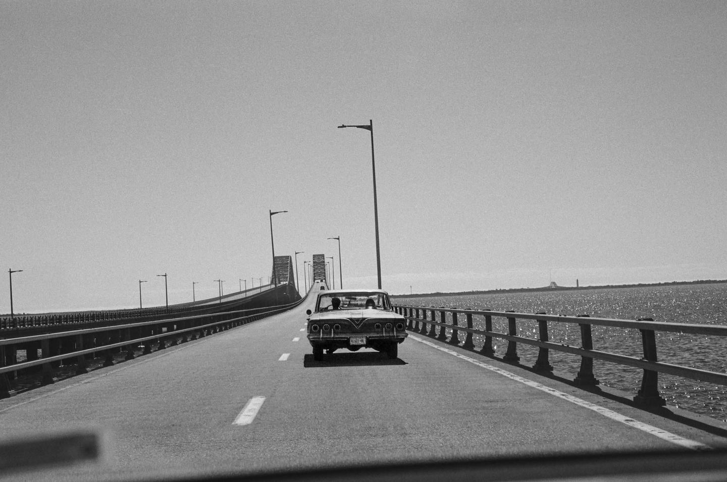 A car drives on a long bridge over water with a clear sky.