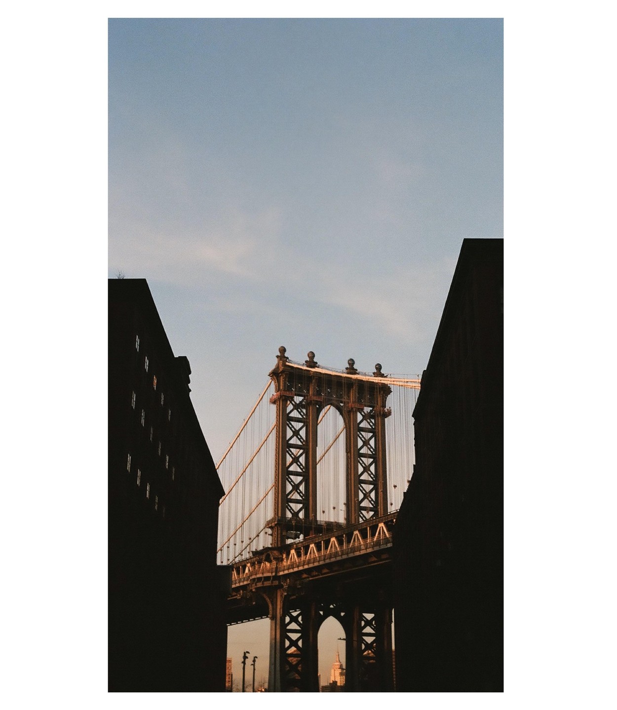 The Manhattan Bridge is framed by two dark buildings against a clear sky.