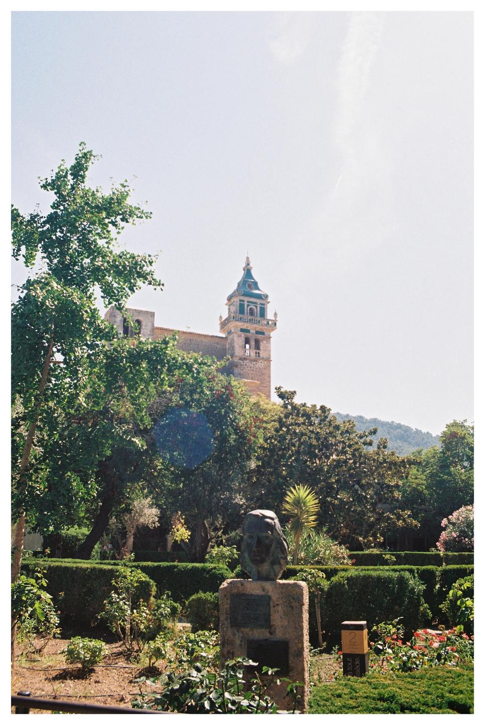 A stone bust on a pedestal stands in a lush garden with a church tower visible in the background.