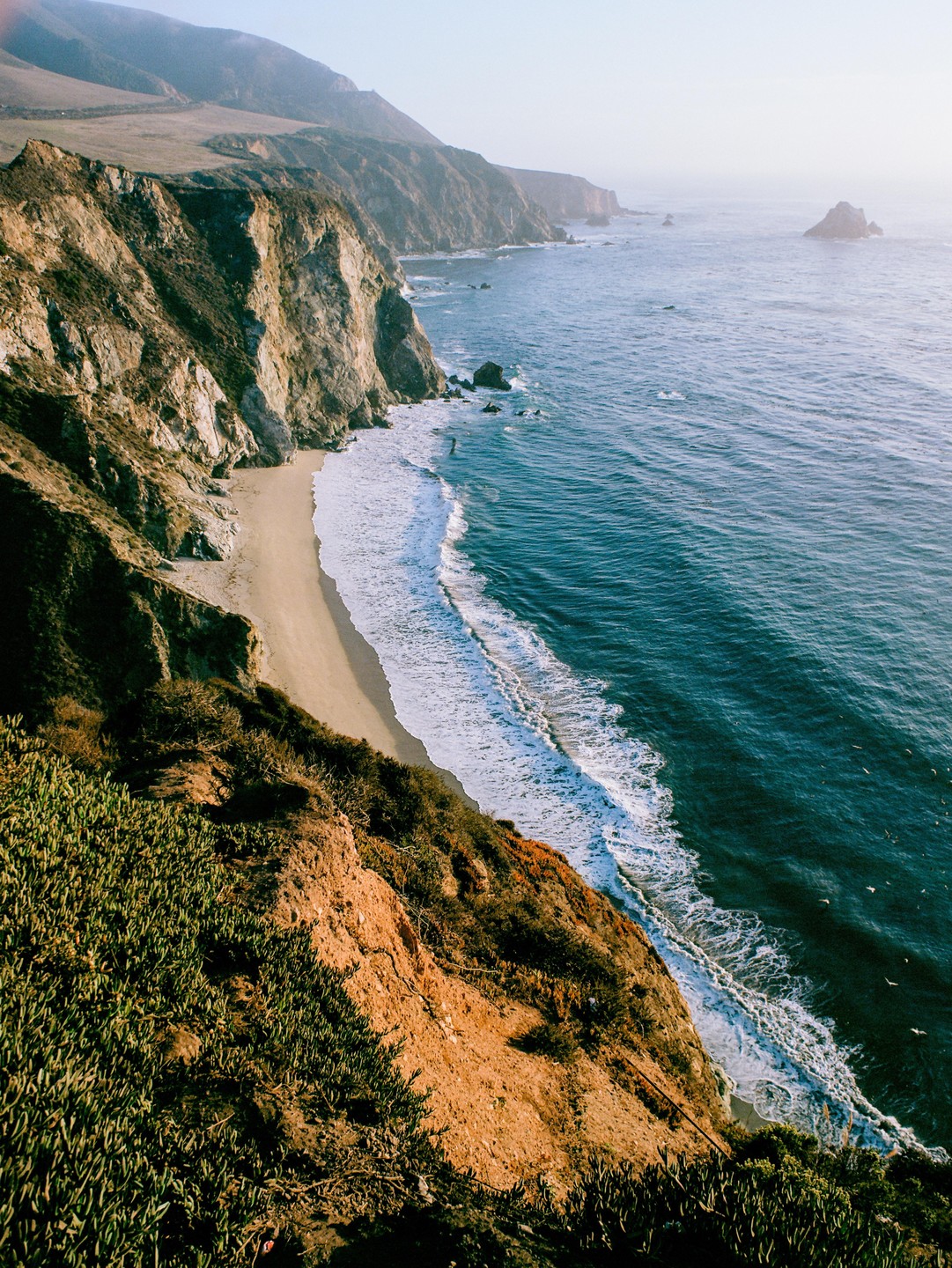Waves crash on a sandy beach at the base of a cliff overlooking the ocean.