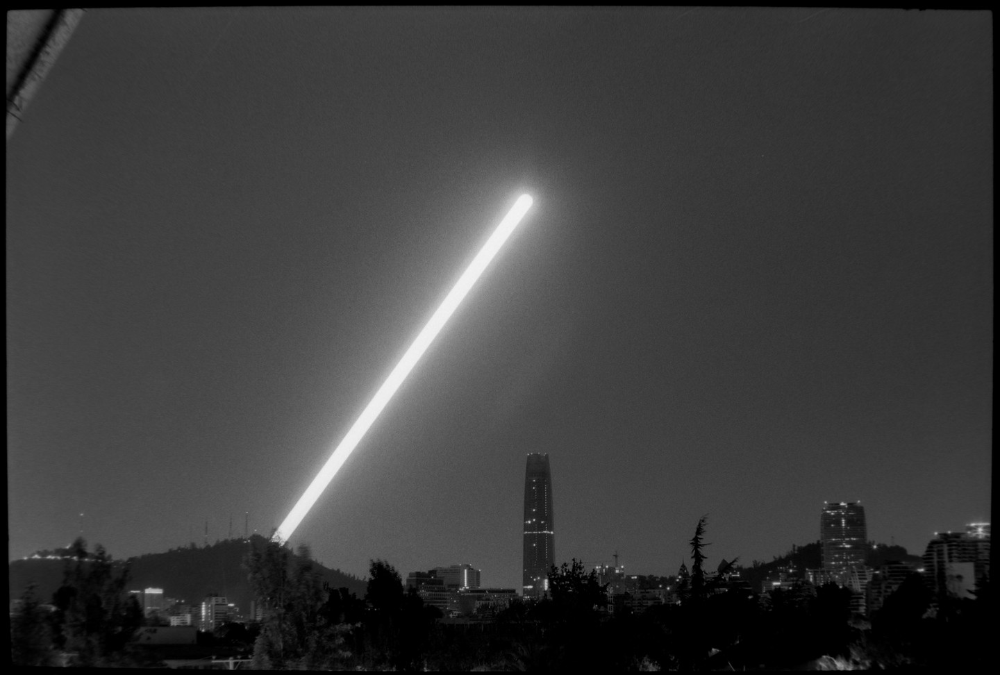A long exposure photograph captures a bright beam of light extending into the night sky above a city skyline with a prominent skyscraper.