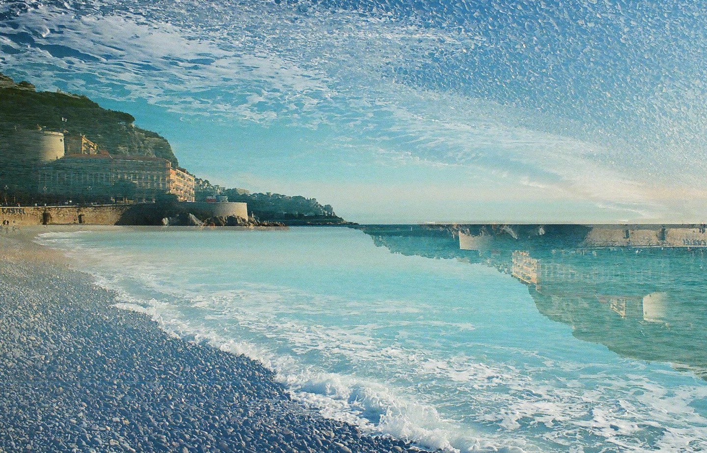 A double exposure shows a pebbled beach with waves crashing on the shore and buildings on a hillside reflected in the water, with a textured sky above.
