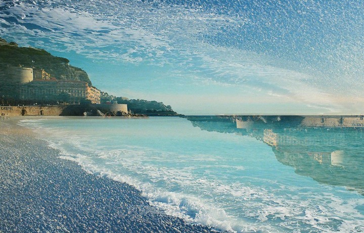 A double exposure shows a pebbled beach with waves crashing on the shore and buildings on a hillside reflected in the water, with a textured sky above.