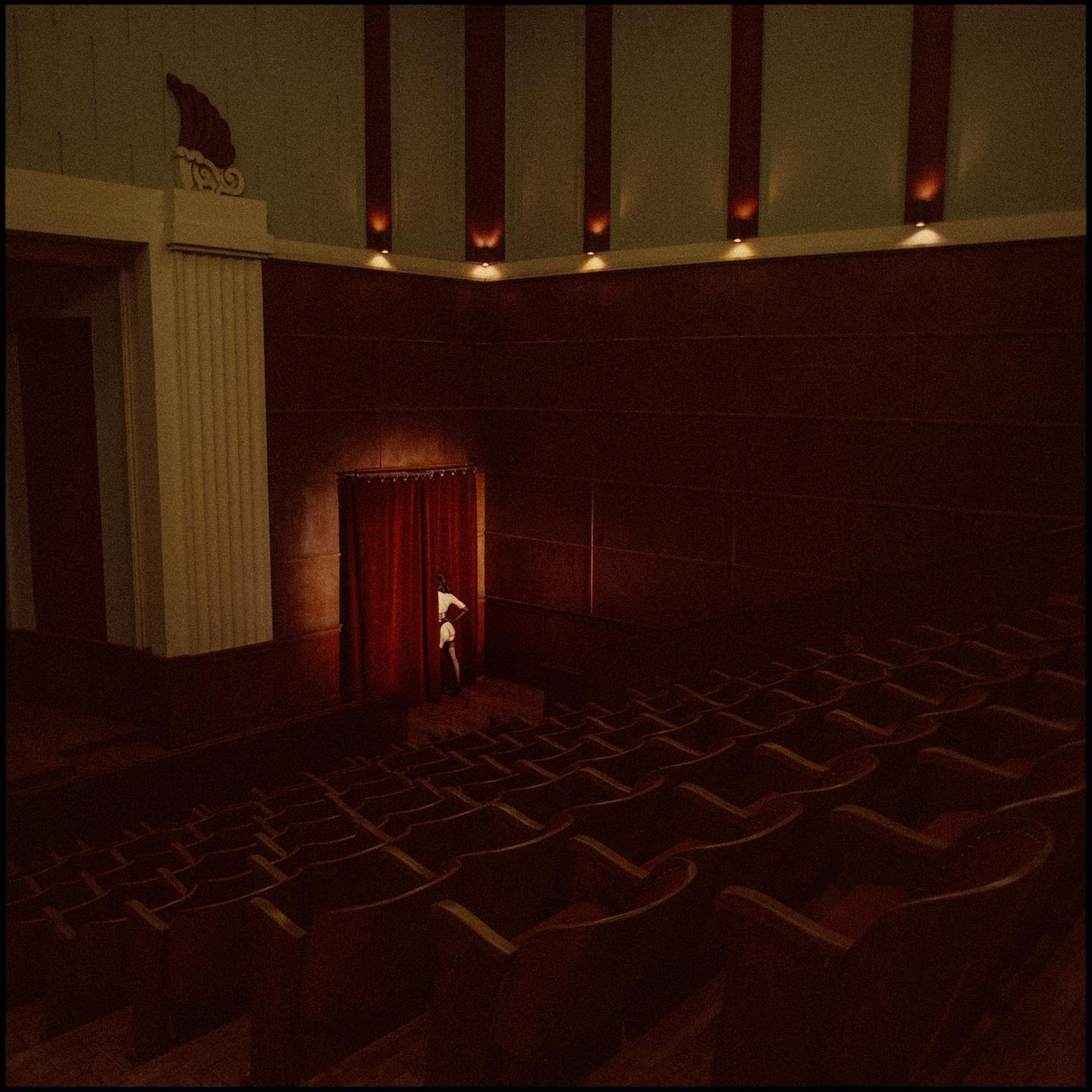 A person peeks from behind a red curtain in an empty theater.