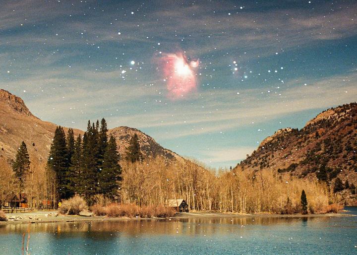 A small cabin sits by a lake surrounded by mountains and trees under a starry sky with a nebula.