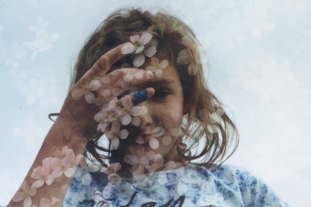 A child with a blue bandage on one finger holds a hand in front of their face, with a double exposure of pink blossoms overlaid on the image.