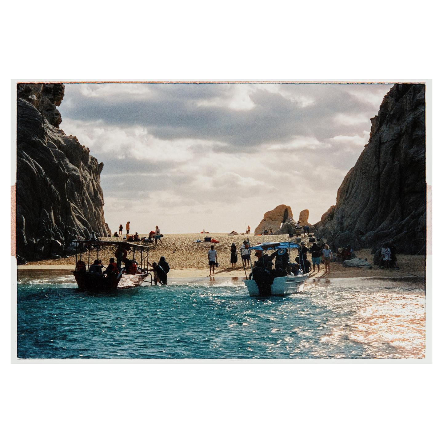 Two boats are on a sandy beach with people and large rock formations under a cloudy sky.