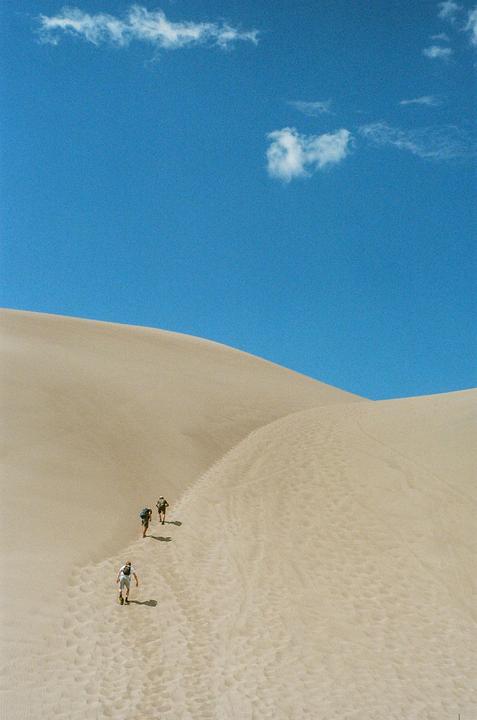 Three people walk up a large sand dune under a clear blue sky with a few clouds.
