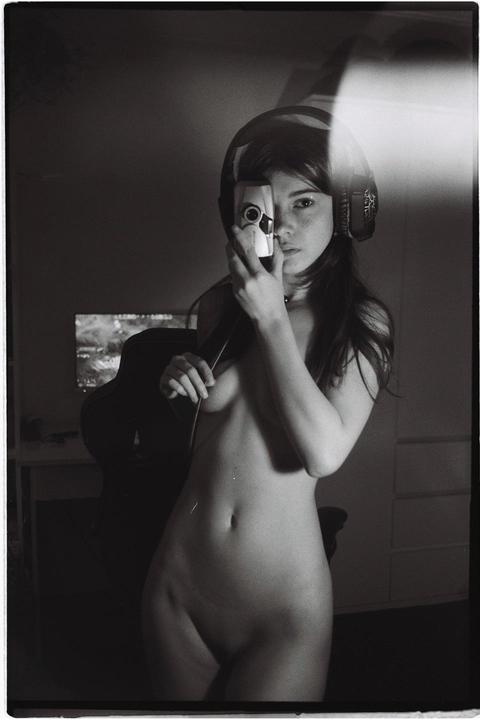 A woman wearing headphones holds a small camera to her face, obscuring one eye, while standing nude in a dimly lit room.