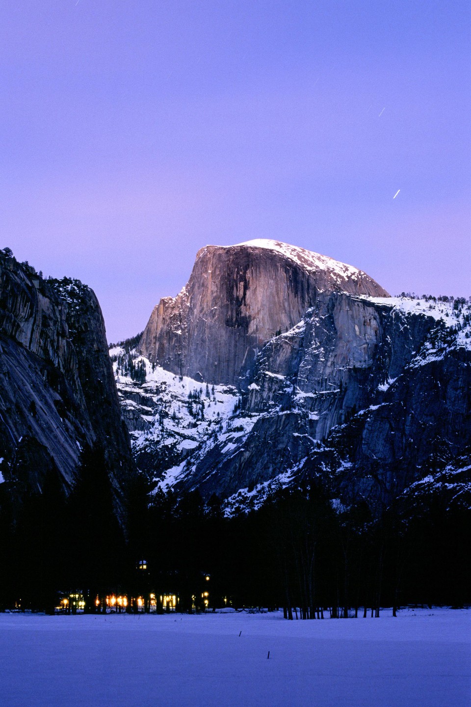 A snow-covered Half Dome mountain rises above a dark forest and illuminated building in a snowy landscape under a purple sky.