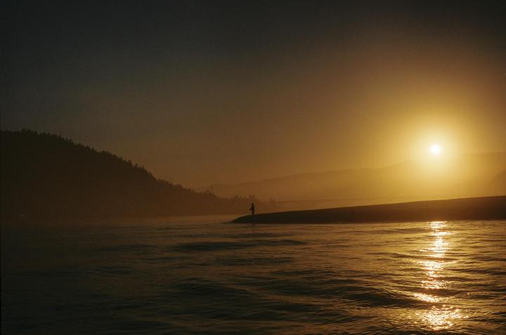 A lone person stands on a narrow strip of land, fishing in a body of water as the sun sets over distant hills.