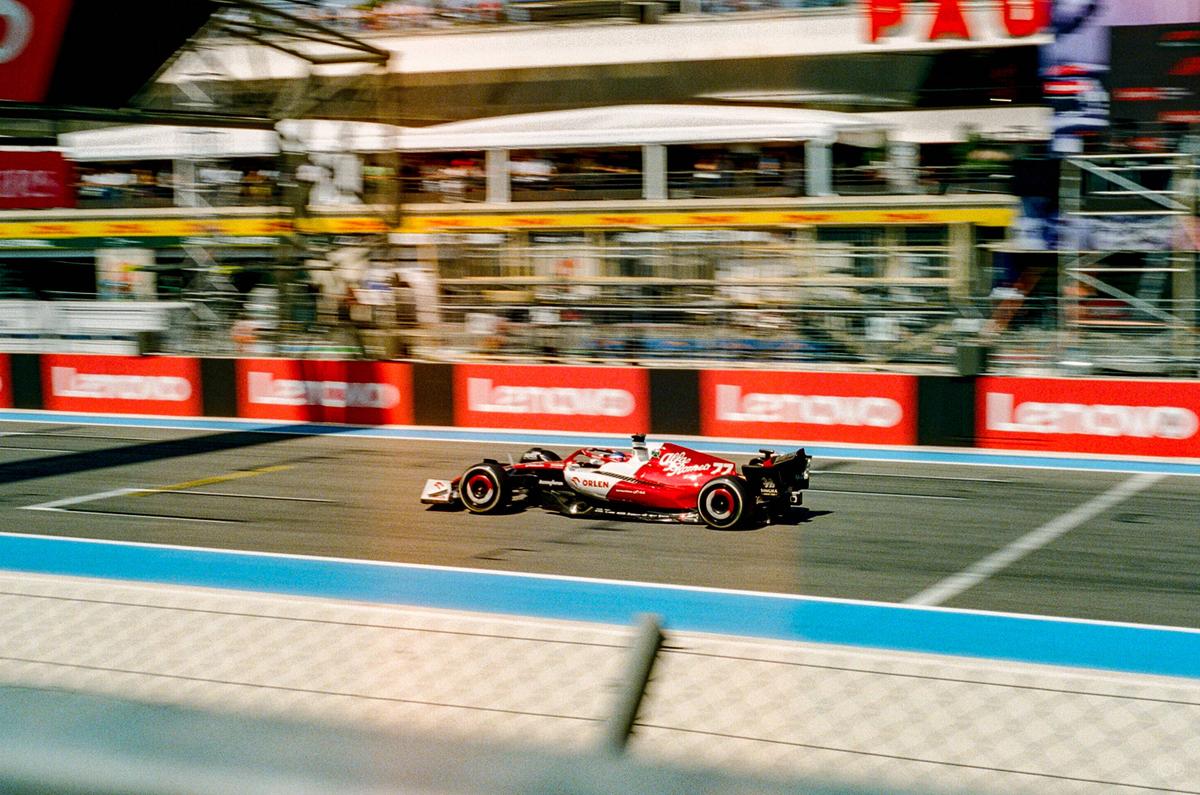 A red and white race car speeds along a track with spectators and buildings in the background.