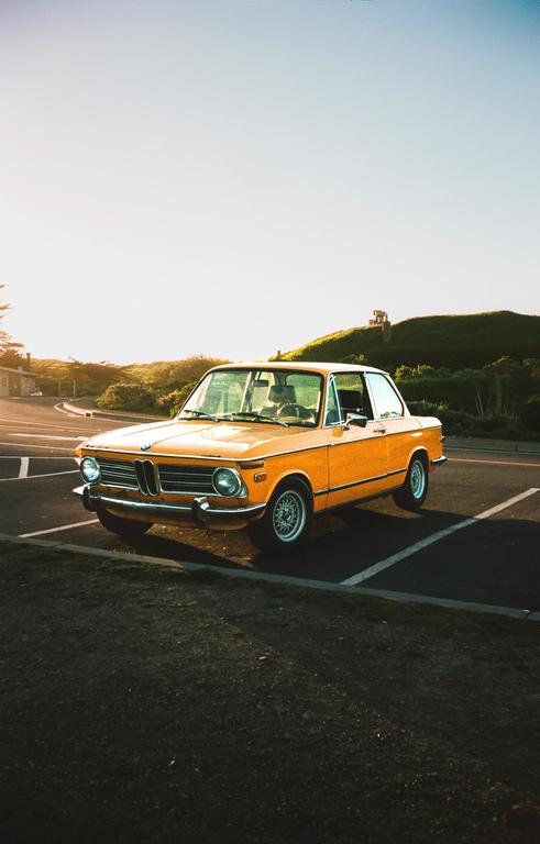 A classic orange car is parked on an asphalt lot with a grassy hill and bright sky in the background.