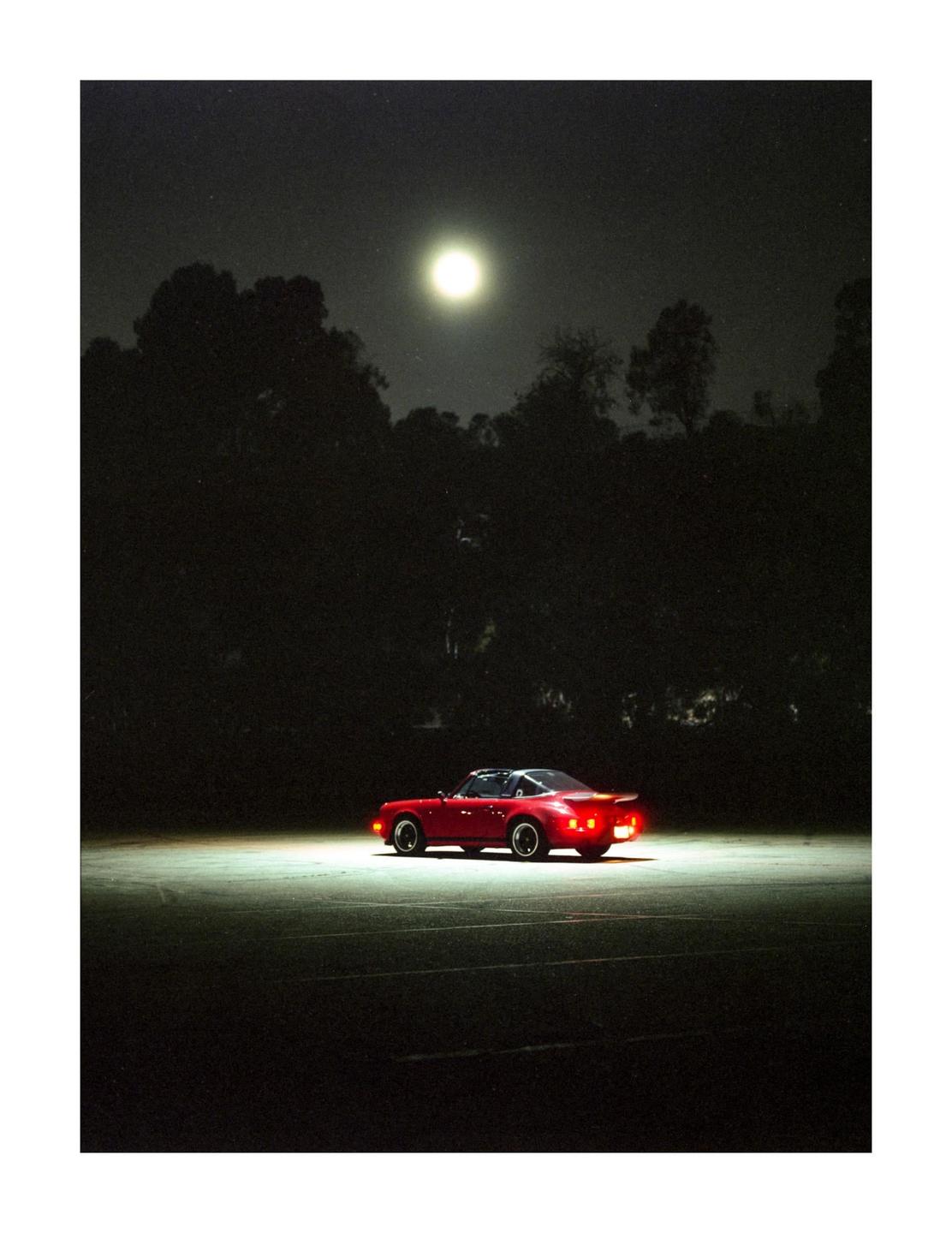 A red sports car is parked on an empty lot under a bright full moon at night.