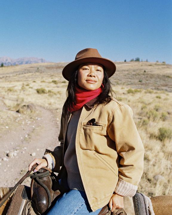 A woman wearing a hat and red scarf sits on a horse in a dry, grassy landscape.