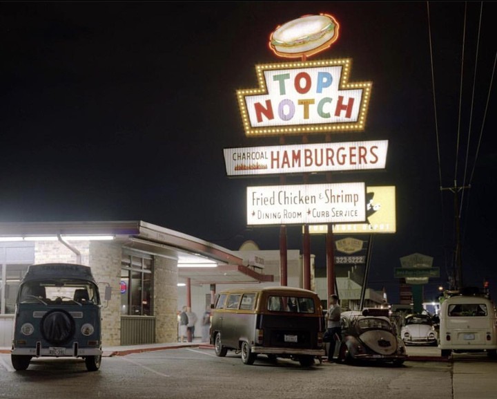 A brightly lit sign for a hamburger restaurant stands over a parking lot full of vans and cars at night.
