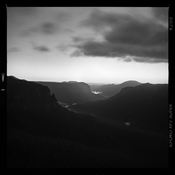 A dramatic black and white landscape shows a valley with mountains and clouds.
