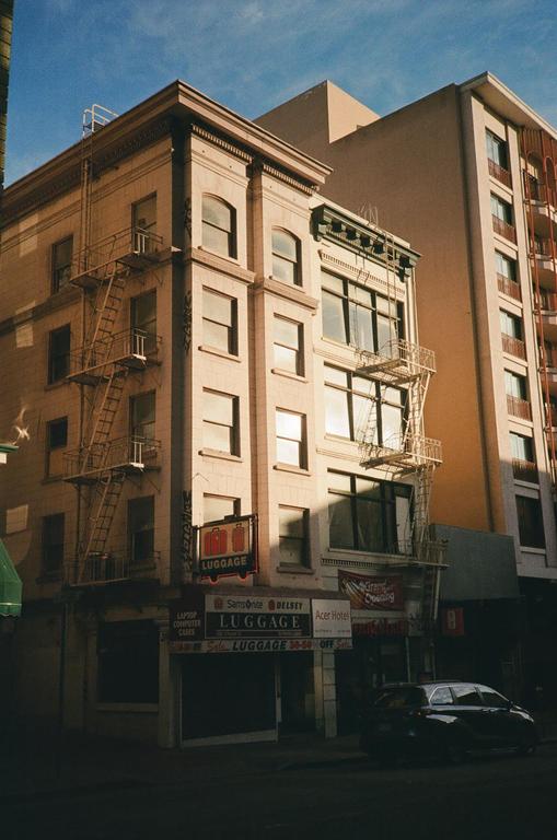 A black car is parked on a street in front of a building with fire escapes and a luggage store.