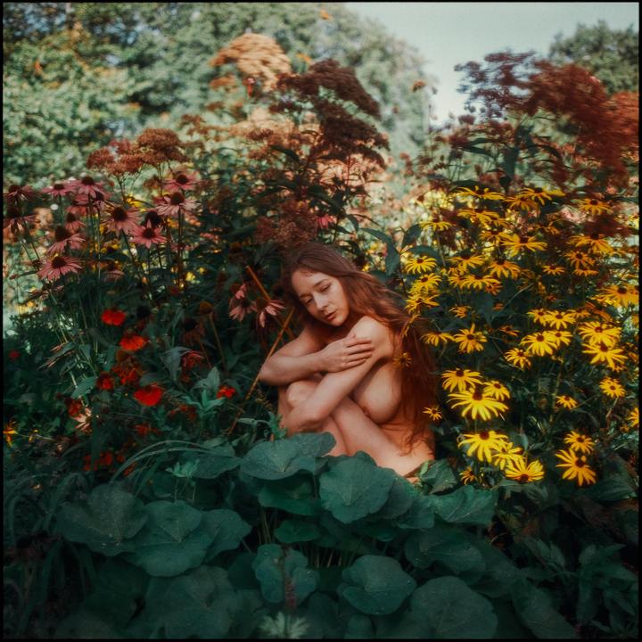 A woman with long hair is sitting nude among colorful flowers and green foliage.