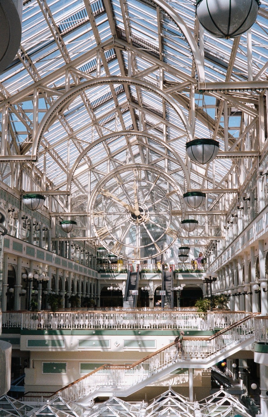 A large, ornate clock hangs from the glass ceiling of a multi-story building with escalators and balconies.