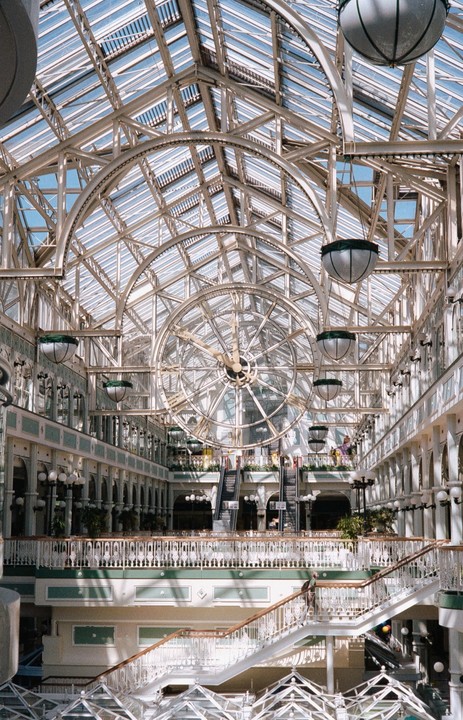 A large, ornate clock hangs from the glass ceiling of a multi-story building with escalators and balconies.