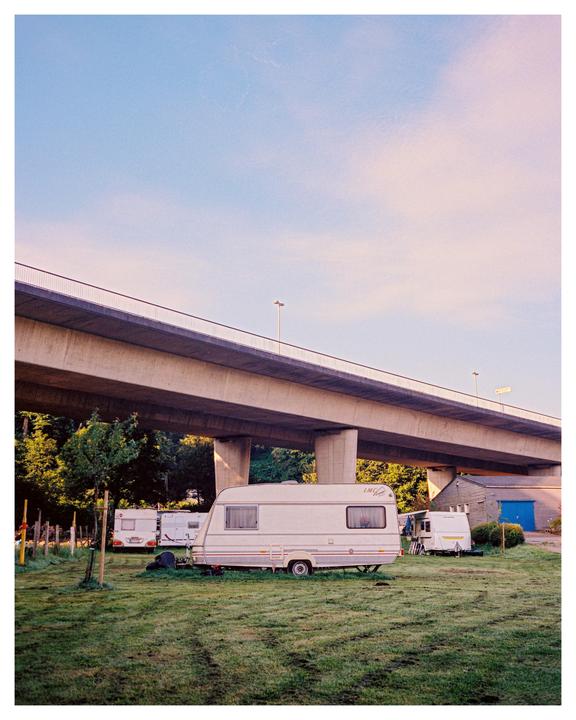 Several caravans are parked on a grassy field under a large concrete overpass.
