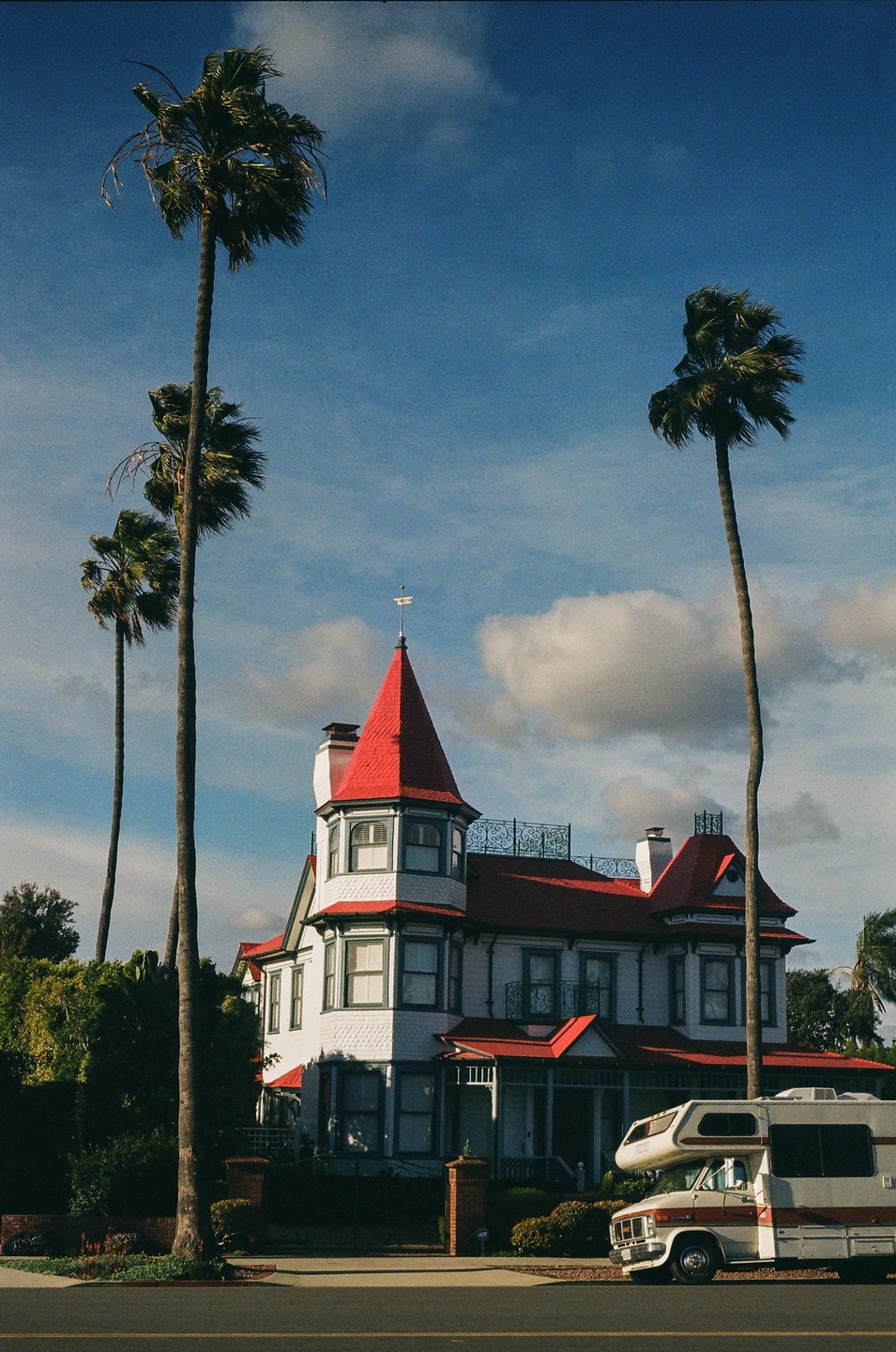 A white Victorian house with a red roof stands between tall palm trees, with a camper van parked on the street in front of it.