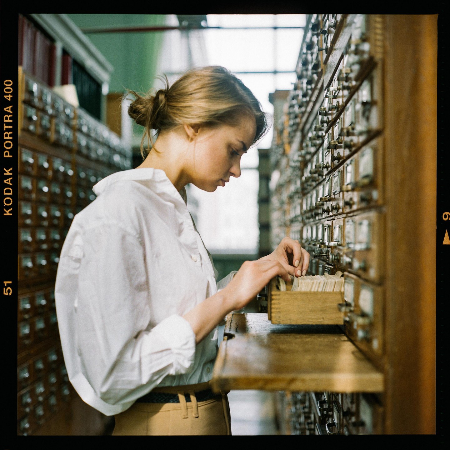A woman looks through index cards in a wooden card catalog in a library.