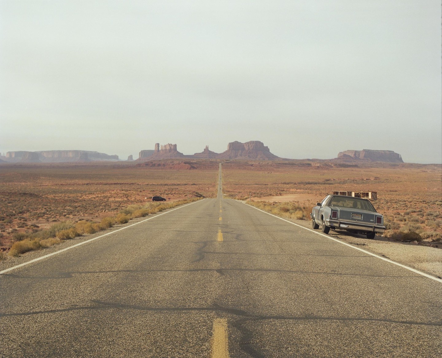 A long, straight road stretches into the distance towards a series of mesas and buttes under a hazy sky, with a car parked on the shoulder in the foreground.