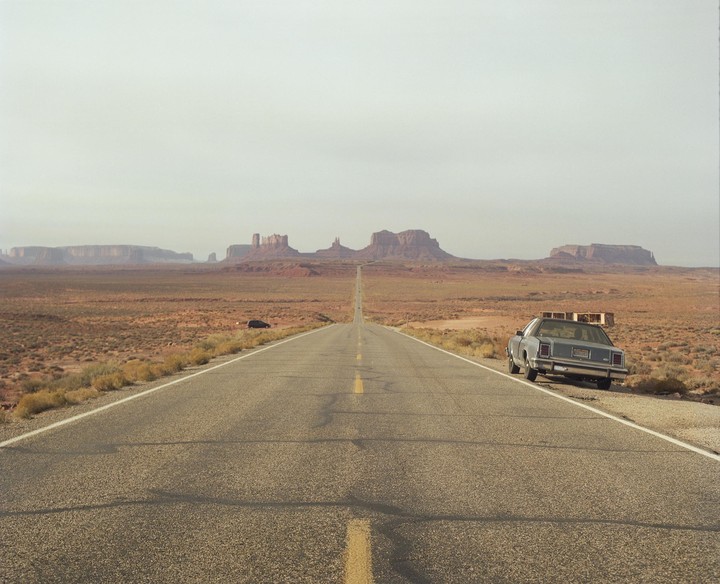 A long, straight road stretches into the distance towards a series of mesas and buttes under a hazy sky, with a car parked on the shoulder in the foreground.