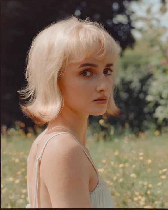 A woman with blonde hair looks over her shoulder in a field of yellow flowers.