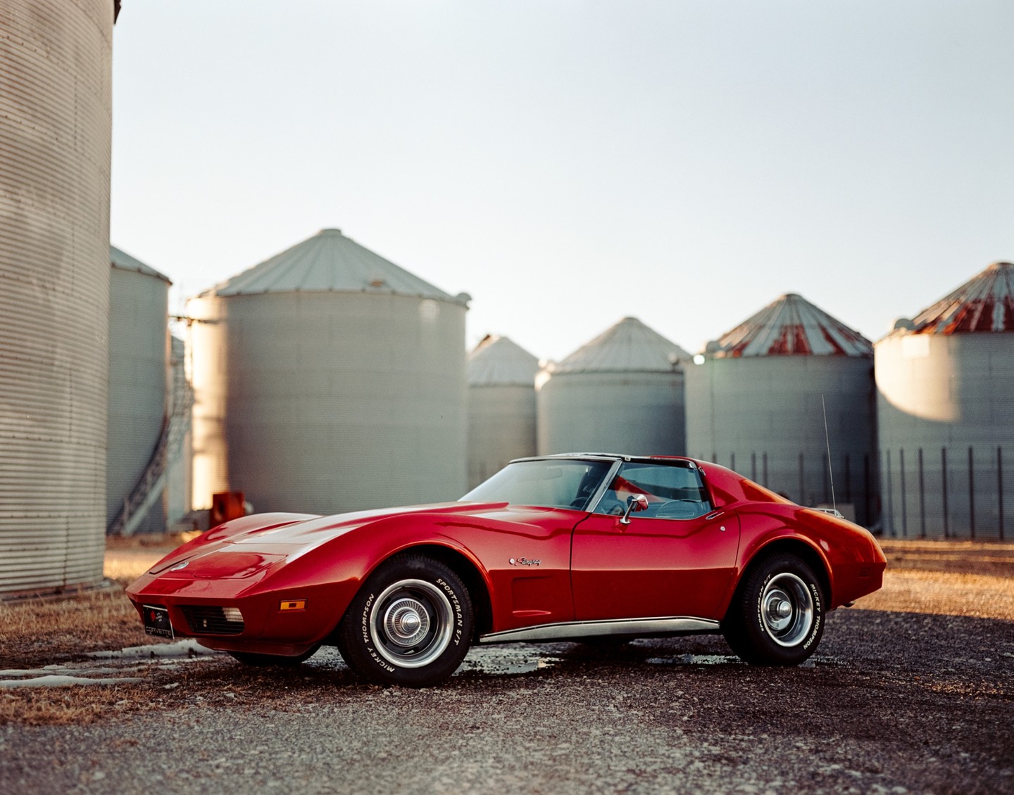 A red Corvette is parked on a gravel road in front of several grain silos.