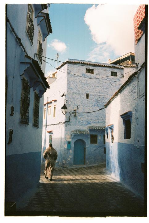 A man in a striped robe walks down a narrow street lined with blue buildings under a cloudy sky.