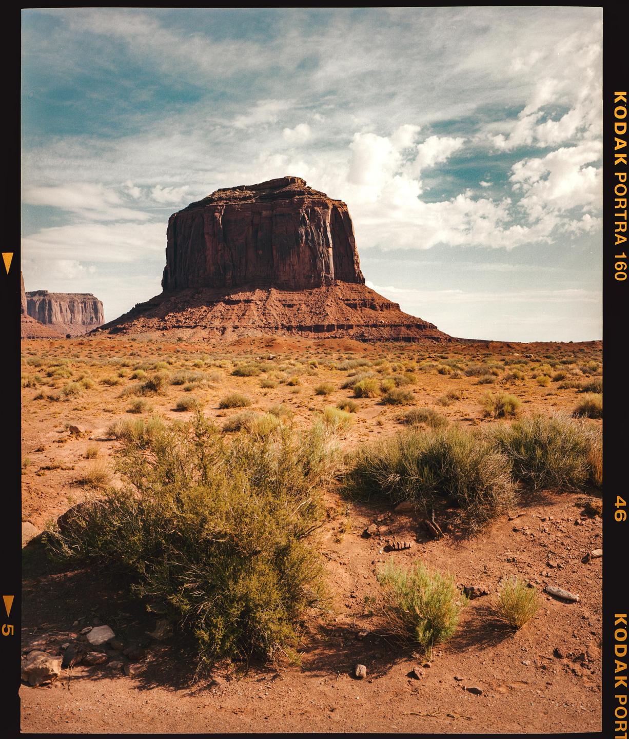 A large red rock formation stands in a desert landscape under a cloudy sky.