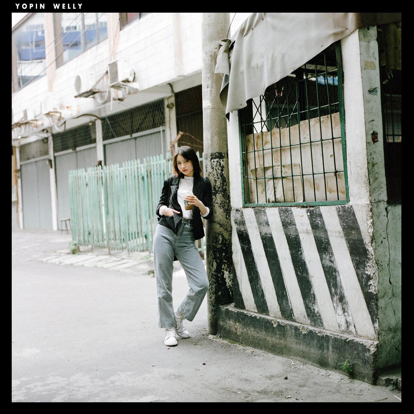 A woman stands on a street holding a drink, leaning against a concrete pillar with black and white stripes.