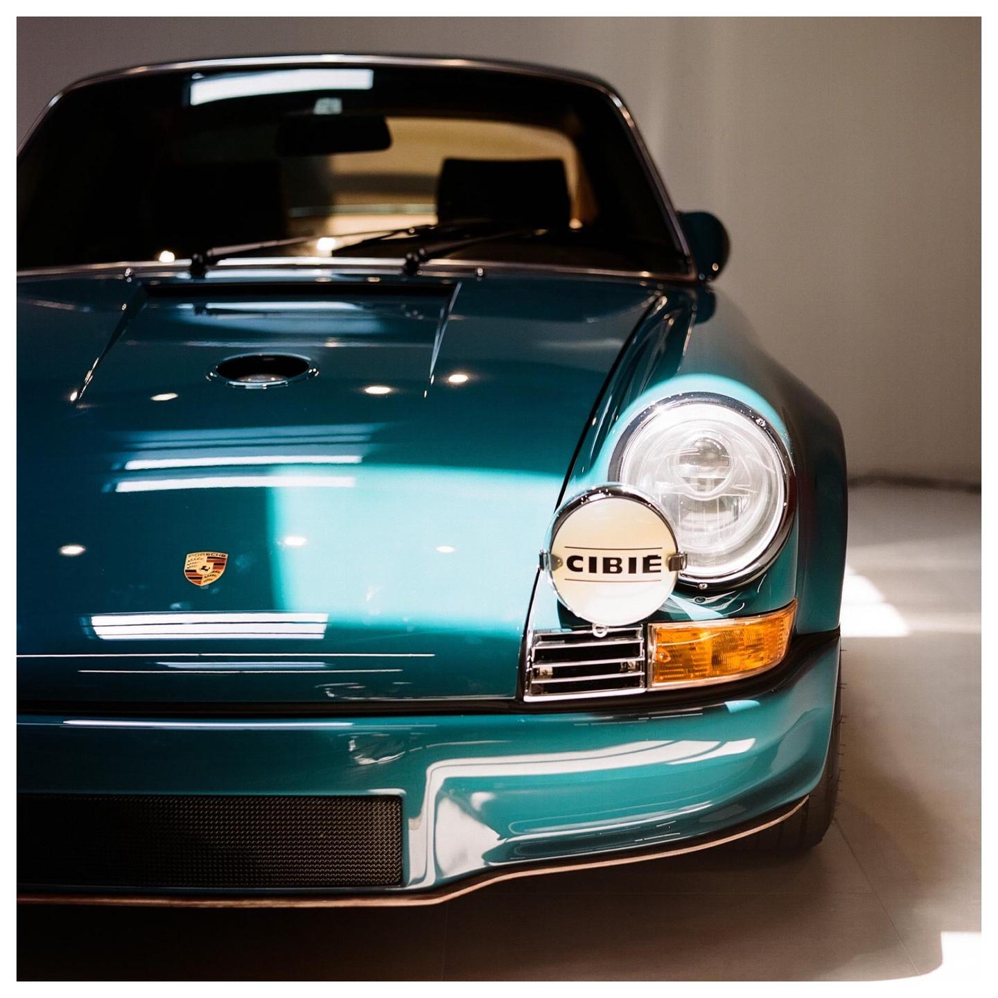 The front of a teal vintage car with a round headlight and a CIBIE auxiliary light is shown.
