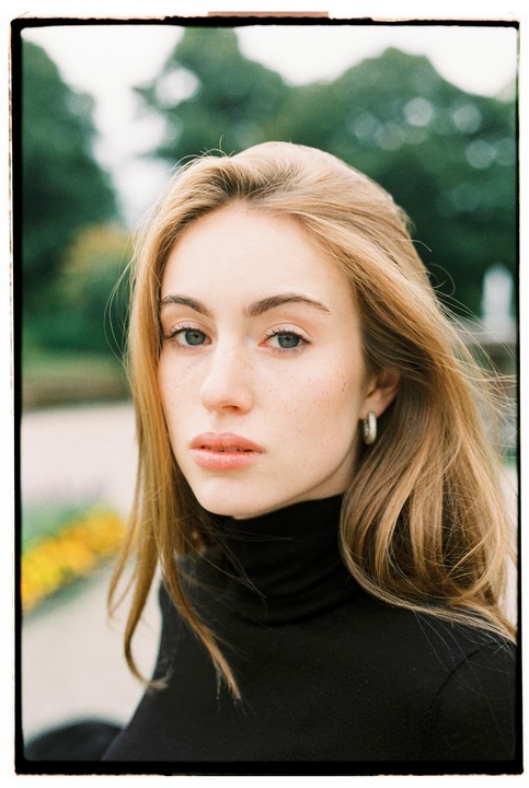 A woman with long hair and a black turtleneck looks directly at the viewer.