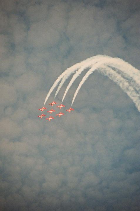 Seven red and white airplanes fly in formation, leaving white smoke trails in a cloudy sky.