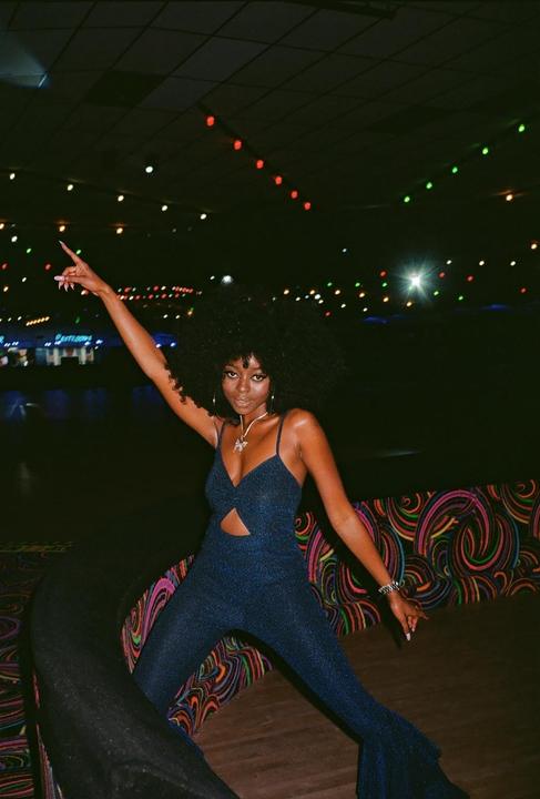A woman in a sparkly blue jumpsuit poses with one arm raised in a roller rink at night.