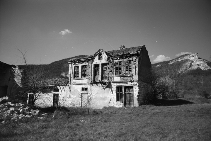 An abandoned house with broken windows and a crumbling facade stands in a grassy field with mountains in the background.